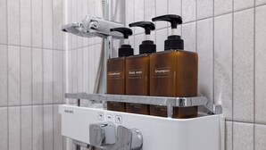 A bathroom with a shower and a shelf holding three bottles of body wash and shampoo. - Grandbleu Hotel & Residence (Incheon)