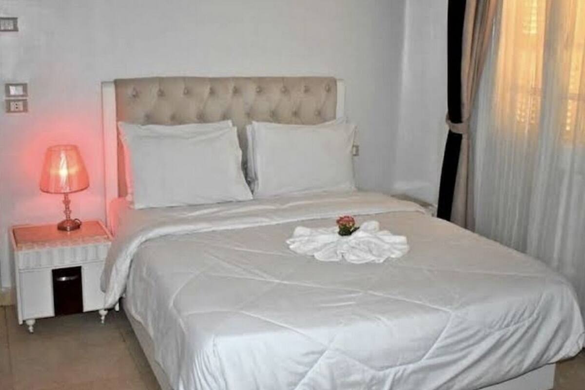 A neatly made bed with a white duvet and pillows, a bedside table with a lamp, and a window with sheer curtains.