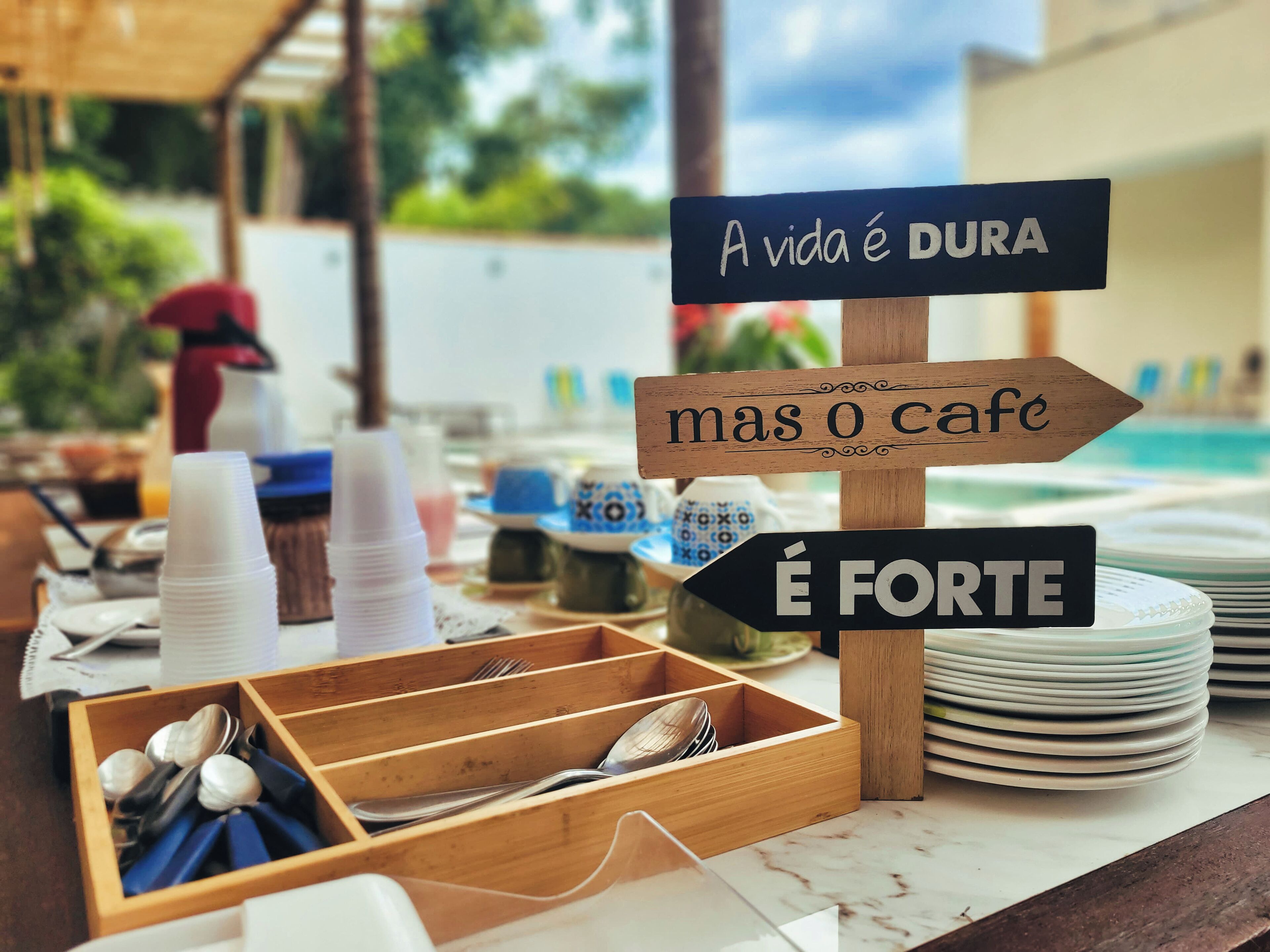 Disposição de buffet com placas em português, uma pilha de pratos e um suporte de utensílios de madeira.