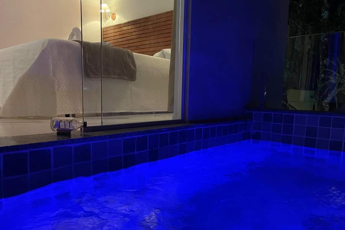 Una habitación con puerta de cristal que da a una piscina interior iluminada en azul, una cama con ropa blanca y un cabecero de madera.