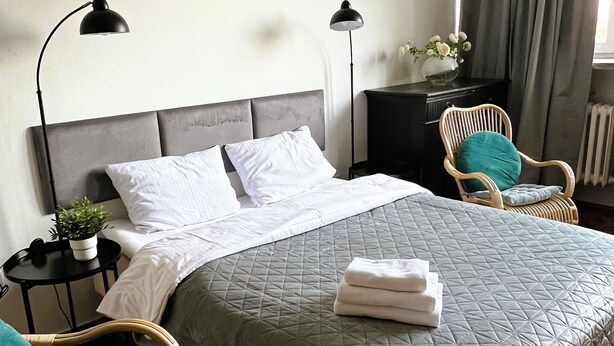A neatly made bed with a grey quilt, two bedside lamps, a small table with a plant, and a wicker chair.