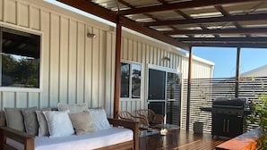 A covered outdoor seating area with a wooden bench and a sofa, a grill, and a view of the surrounding area.