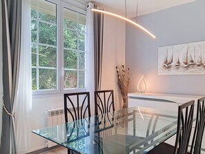 A modern dining area with a glass table, chairs, a radiator, a window with curtains, and a painting of sailboats.