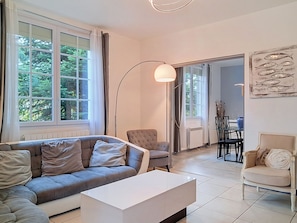 Una sala moderna con un sofá gris, una mesa de centro blanca y una lámpara de pie. Se ve un área de comedor con una mesa y sillas a través de una puerta abierta.