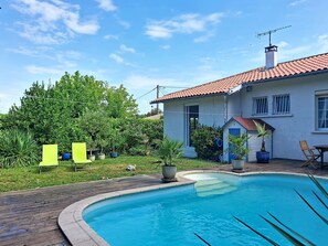 Uma piscina com deck de madeira, uma casa com telhado ladrilhado em vermelho e folhagem verde ao fundo.