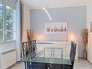 Un espace repas moderne avec une table en verre, des chaises noires, un radiateur blanc, un tableau représentant des voiliers et un vase contenant des plantes séchées.