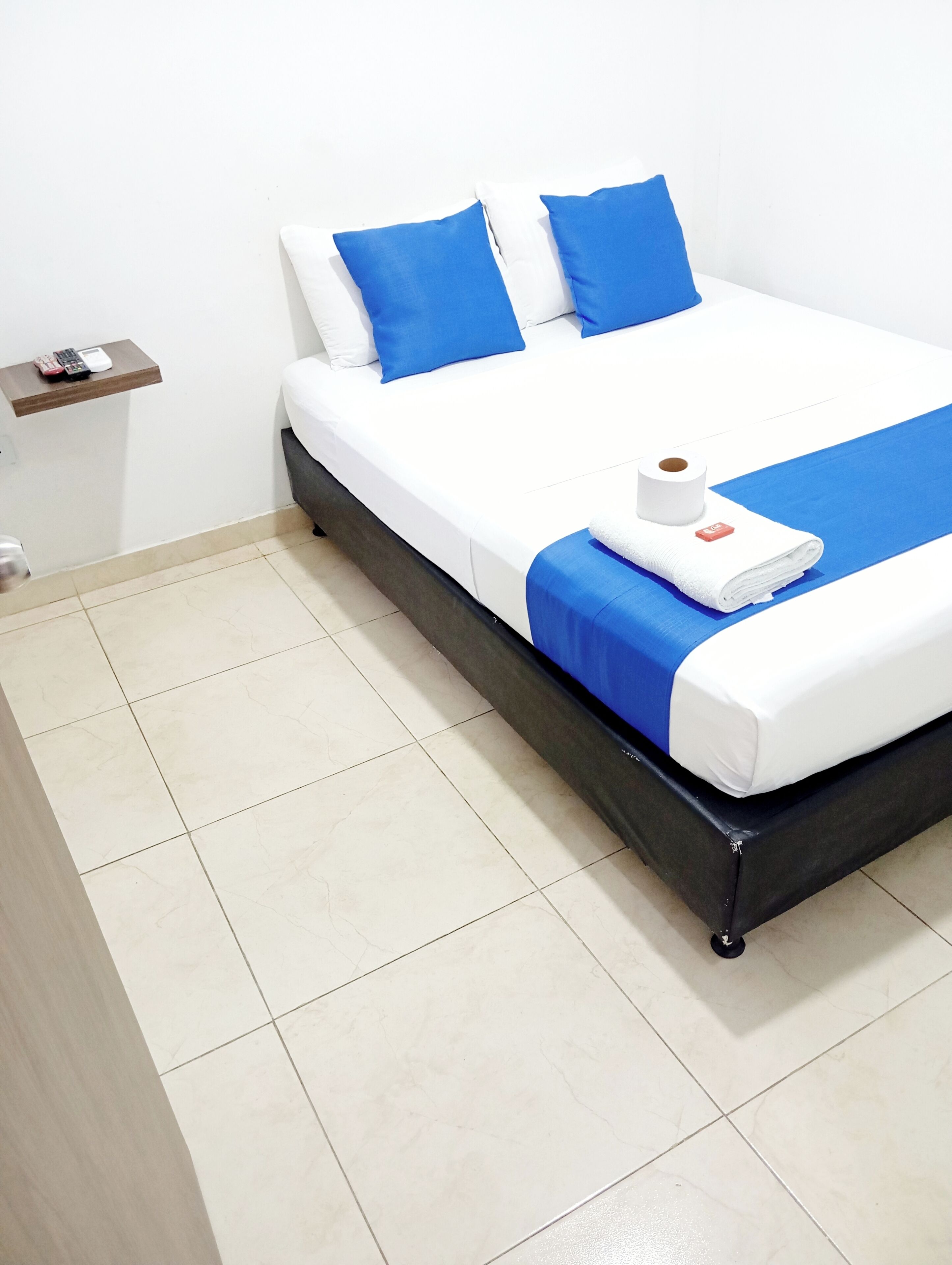 Een opgemaakt bed met blauw-wit beddengoed, een rol toiletpapier en een klein plankje met een telefoon.