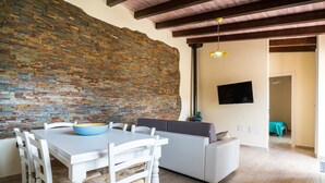 Un comedor con pared de ladrillo, una mesa blanca con sillas, un sofá gris y un televisor montado en la pared.
