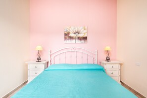 A bed with a teal bedspread, two bedside tables with lamps, a white metal headboard, and a pink wall with a floral painting.