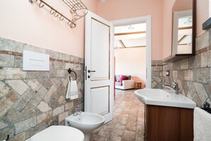 Une salle de bain avec un mur en pierre, un lavabo blanc, un bidet et une porte donnant sur l’espace de vie.