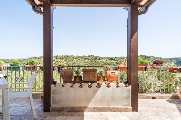 Un espace extérieur abrité offrant une vue sur un paysage vallonné, avec une chaise blanche et des jardinières en terre cuite.