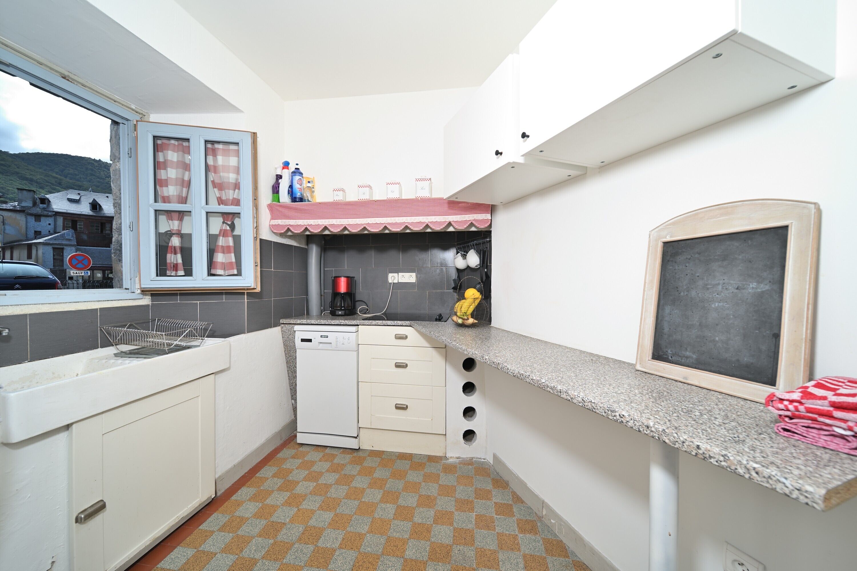 A compact kitchen with a dishwasher, a small sink, a window with checkered curtains, a chalkboard, and a tiled floor.