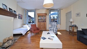 A room with a bed, a white coffee table with books, a red armchair, a plaid armchair, and a view of a mountain through open windows. - Characterful House in Arreau – Mountain View, Balcony, WiFi, Free Parking, 6 Guests, Pets Welcome (Arreau)