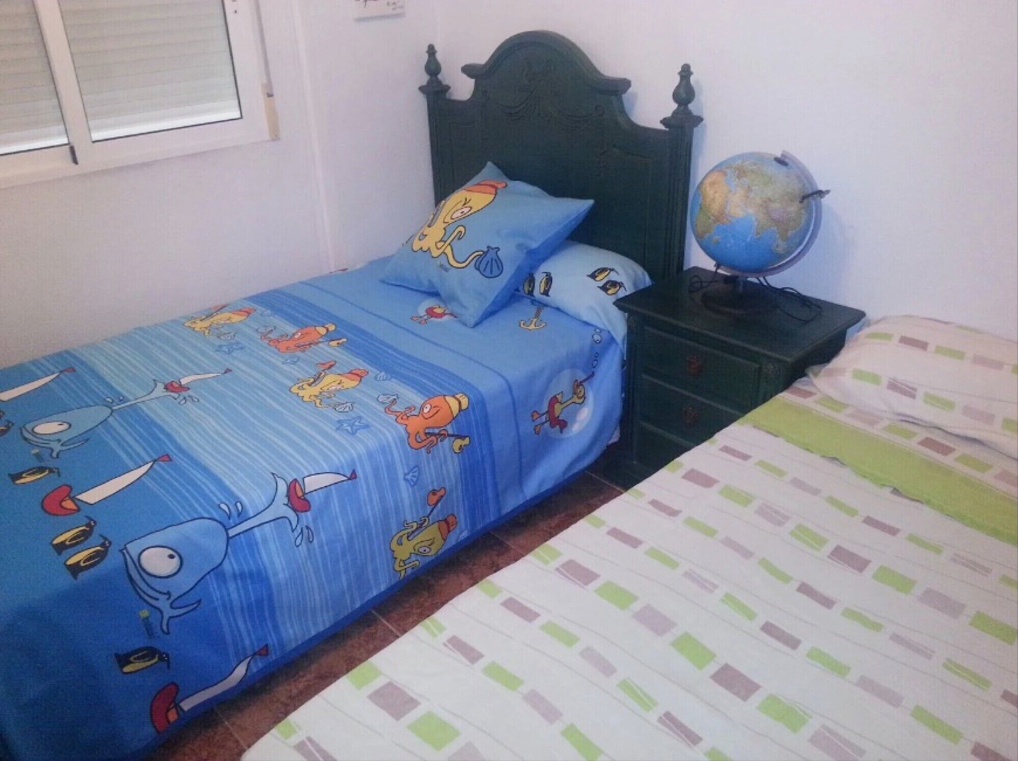 Een slaapkamer met een bed met een blauwe sprei met tekenfilmfiguren, een houten hoofdbord, een nachtkastje met een wereldbol en een raam met luiken.