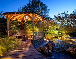 A wooden gazebo with a lattice railing, a wooden bench, and a small pond with rocks.