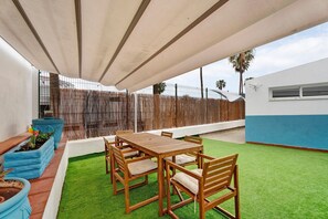 Un patio con mesa y sillas de madera, rodeado de césped artificial, bajo una zona sombreada.