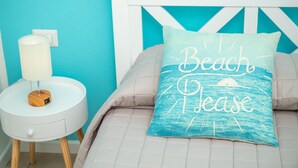 Un lit avec un oreiller sur lequel est écrit « Plage, s’il vous plaît », une table de chevet blanche avec une lampe, et un mur bleu.