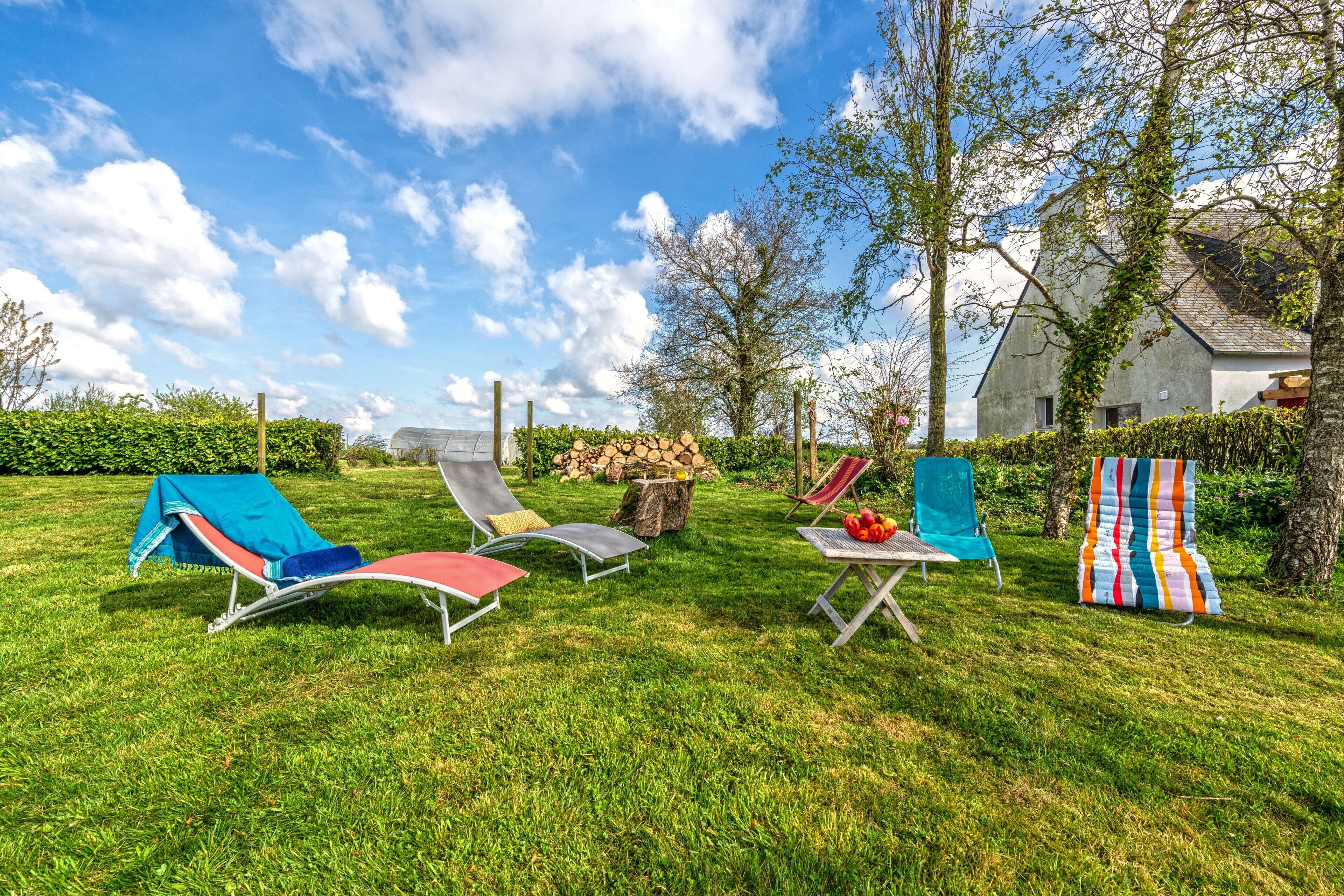 Un espace extérieur verdoyant avec des chaises longues, une petite table sur laquelle se trouve un panier, et un tissu rayé aux couleurs vives.