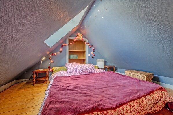 A bedroom with a sloped ceiling, a bed with a red blanket, a bedside table, a lamp, a shelf with books, and a basket.