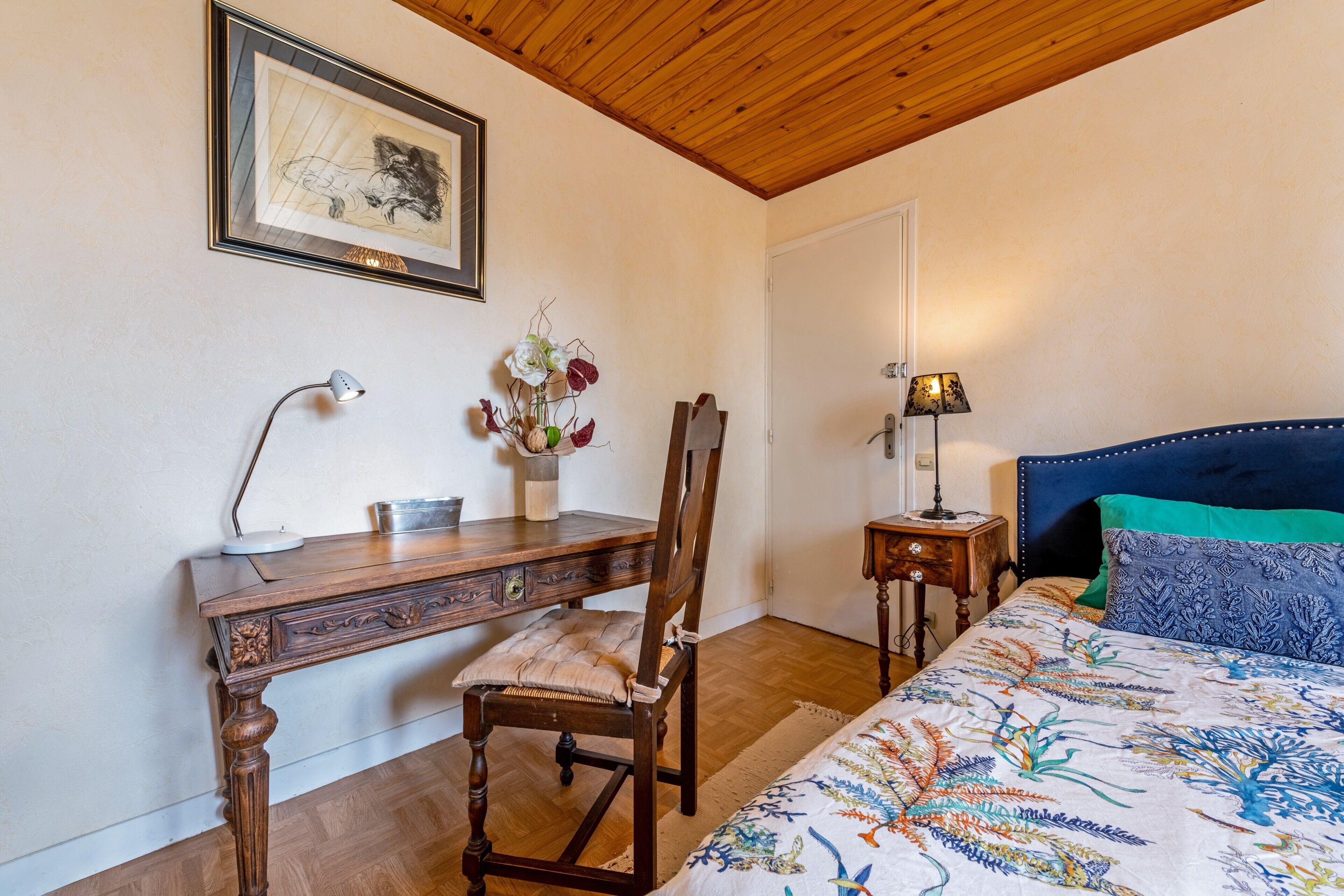 Une chambre à coucher avec un bureau en bois, une chaise, une table de chevet, une lampe et un lit recouvert d’une couvre-lit à motifs.