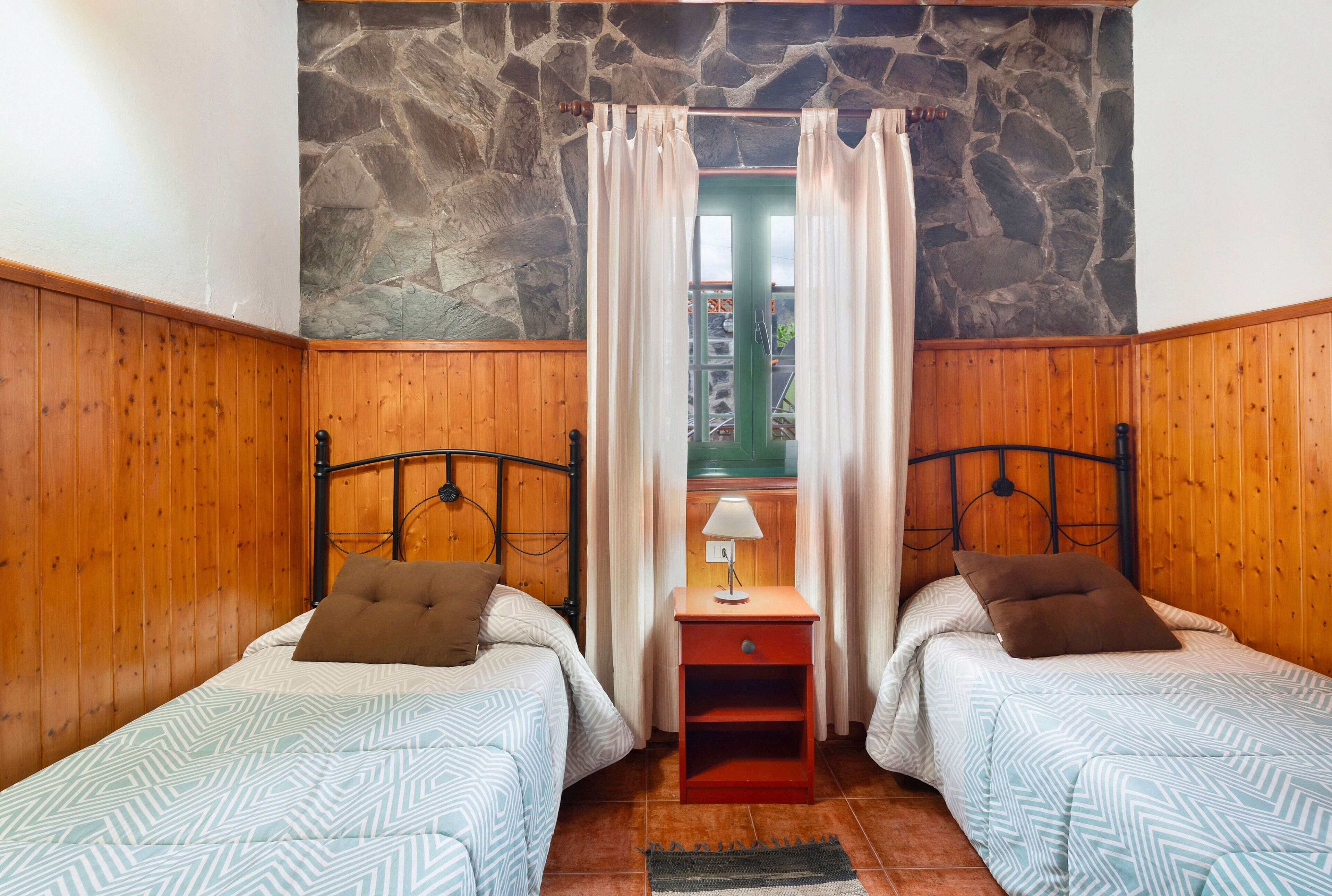 Une chambre avec deux lits, un mur en pierre, un lambris en bois, une table de chevet avec une lampe et une fenêtre avec des rideaux.