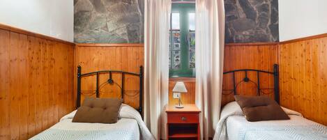Une chambre avec deux lits, un mur en pierre, un lambris en bois, une table de chevet avec une lampe et une fenêtre avec des rideaux.