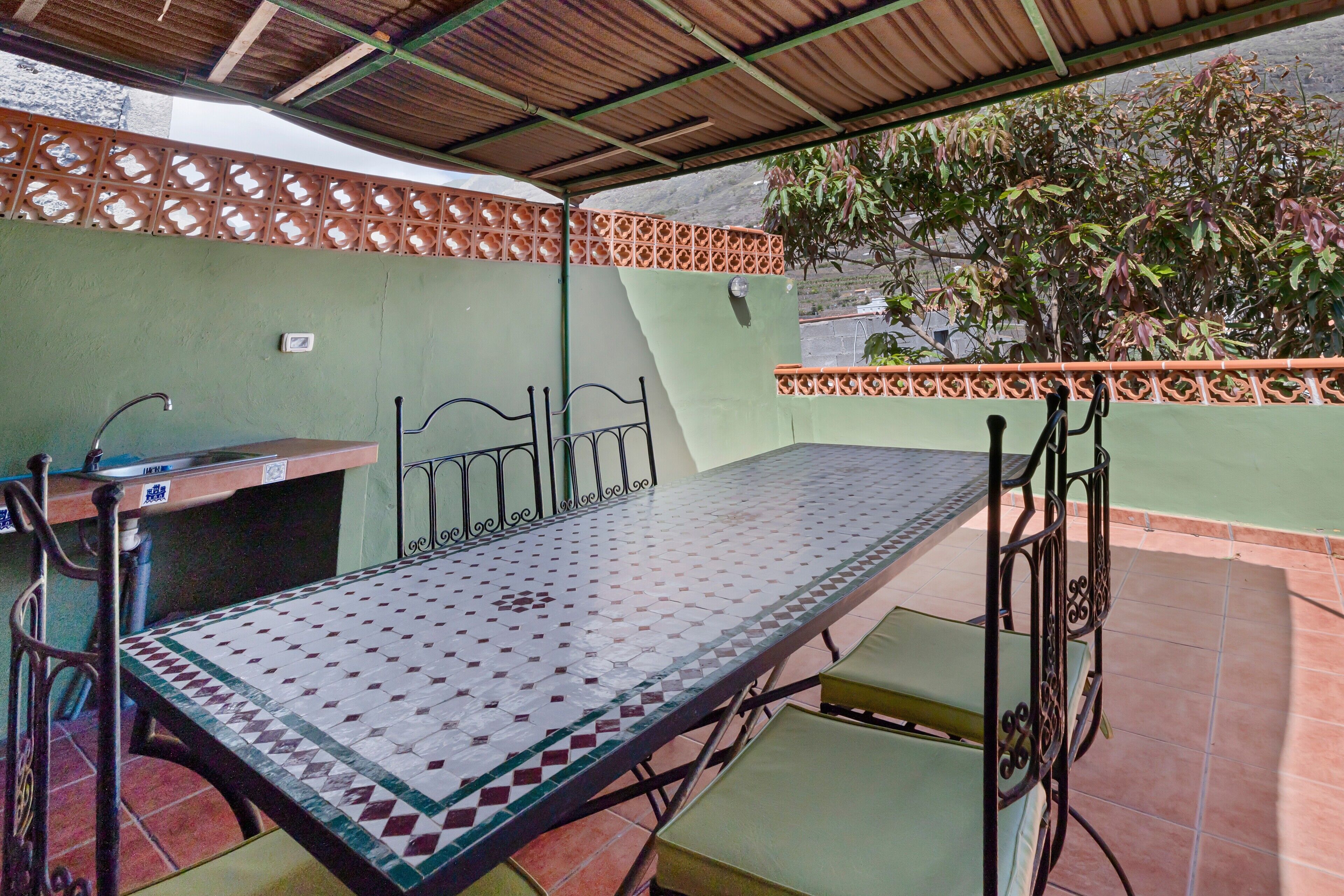 A covered outdoor patio with a metal table and chairs, a sink, and a green wall with a decorative fence.