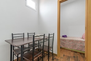 Una habitación con una mesa de comedor de madera, cuatro sillas, una cama pequeña con colcha estampada y una puerta de madera.