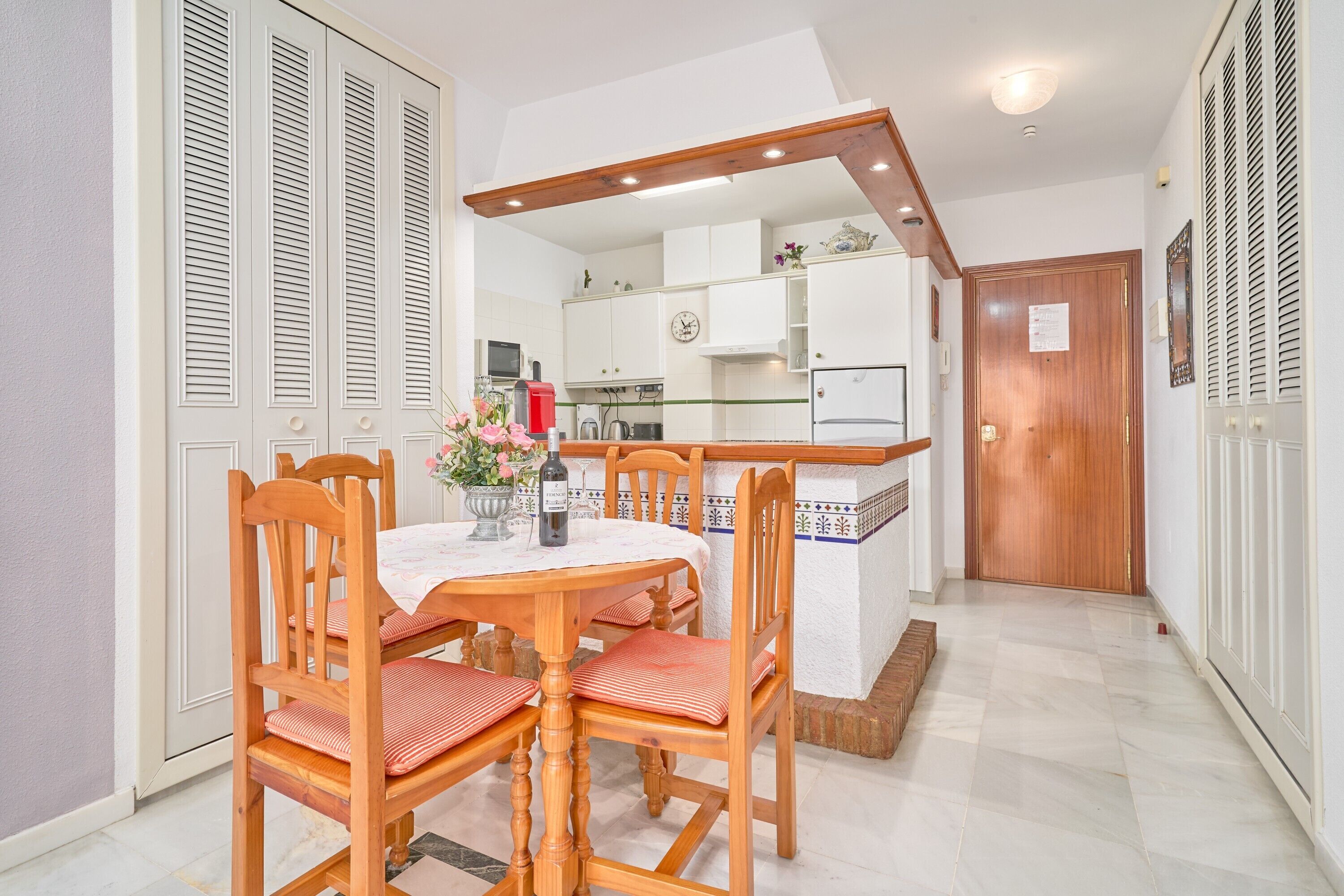 A dining area with a wooden table and chairs, a kitchen in the background, and a wooden door.