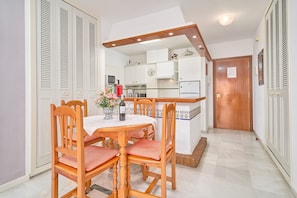 A dining area with a wooden table and chairs, a kitchen in the background, and a wooden door.