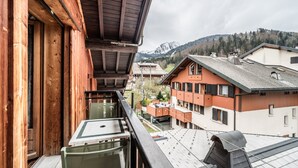 A balcony with a table and chairs, a wooden building, and a view of other buildings and mountains in the background.