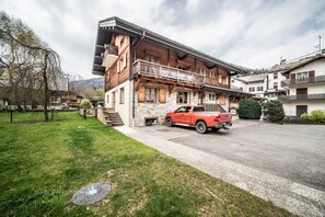 Une maison en bois à deux étages avec un balcon, un camion rouge garé et une cour verdoyante.