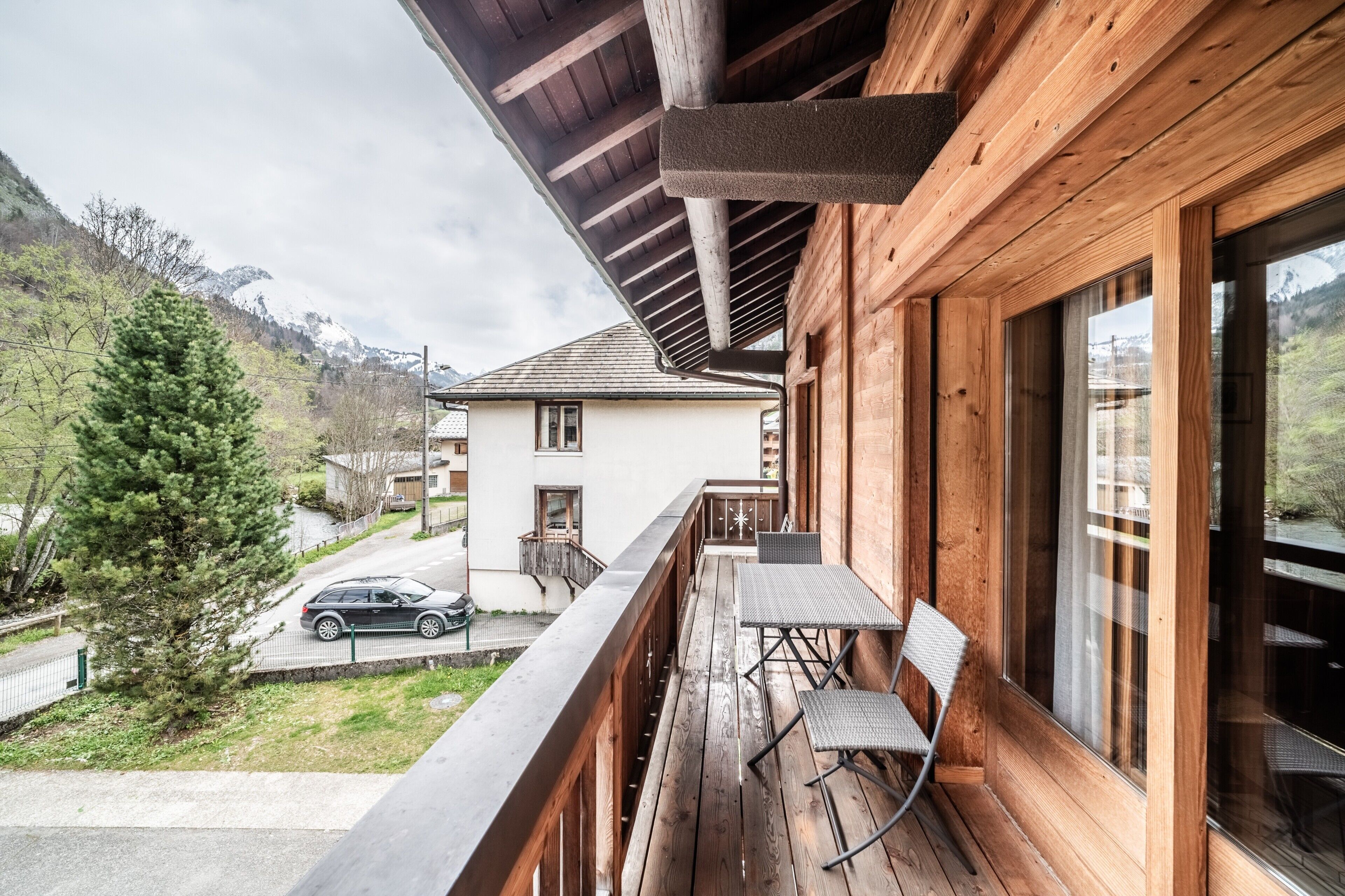 A wooden balcony with a table and chairs, overlooking a residential street and a mountain.