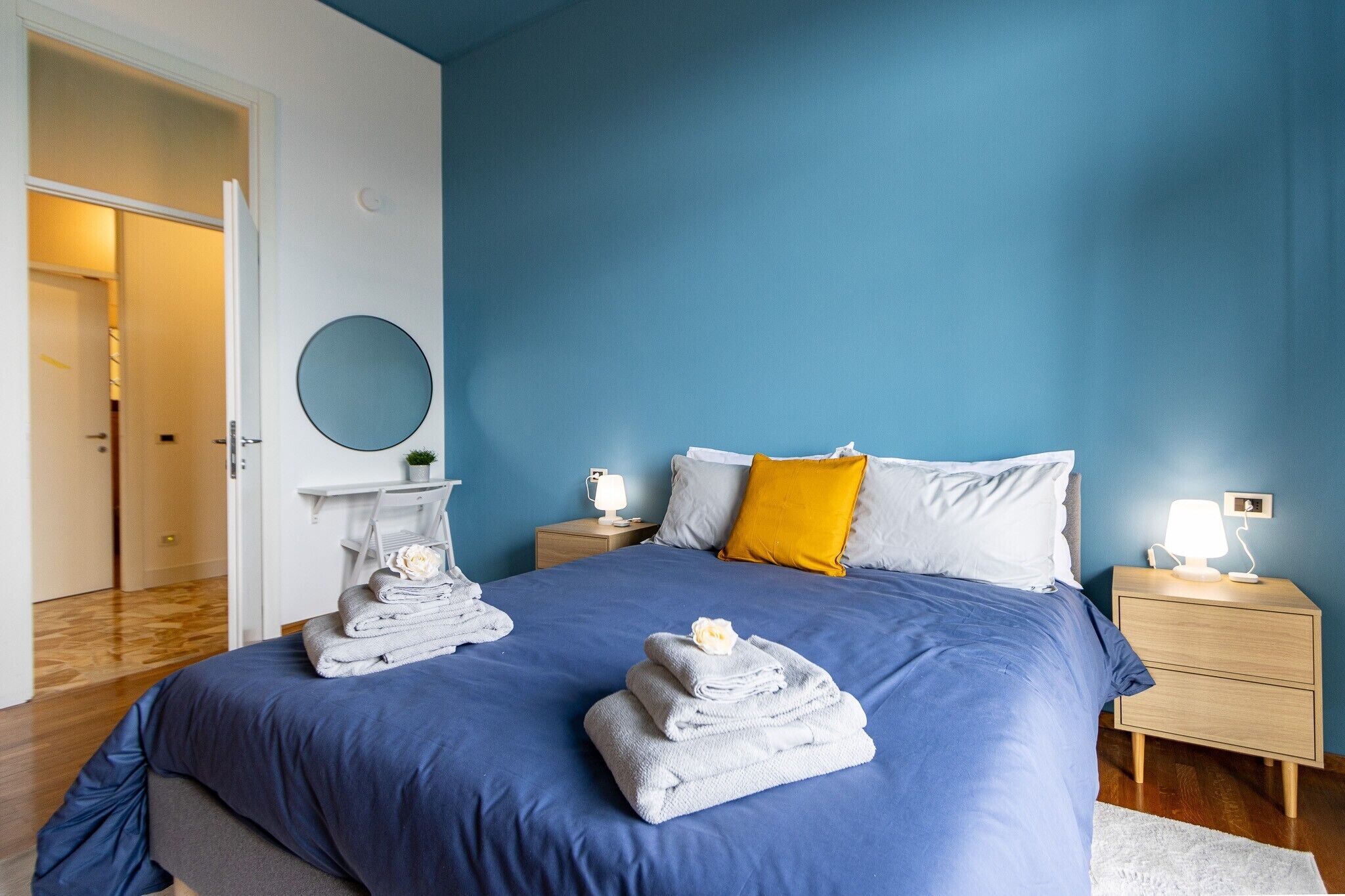 A bedroom with a blue bedspread, a round mirror, a white nightstand with a lamp, and a wooden floor.