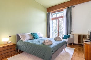 A bedroom with a bed, bedside table, lamp, chair, and a view of the outdoors.