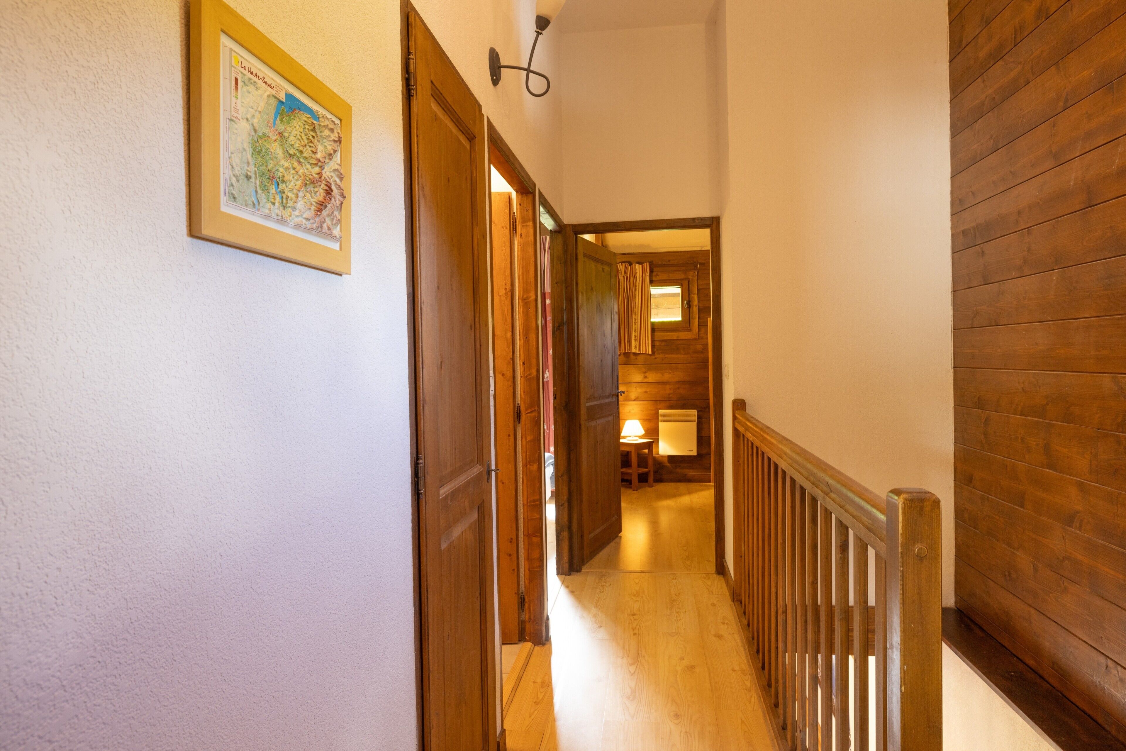 Un couloir avec des portes en bois, une carte encadrée et une lampe visible au loin.