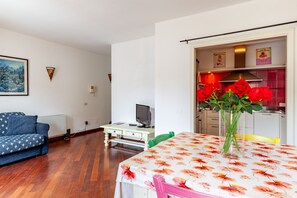 Una sala de estar con un sofá, una mesa de comedor con mantel floreado, un televisor sobre un mueble bajo, y una cocina visible a través de una puerta abierta.