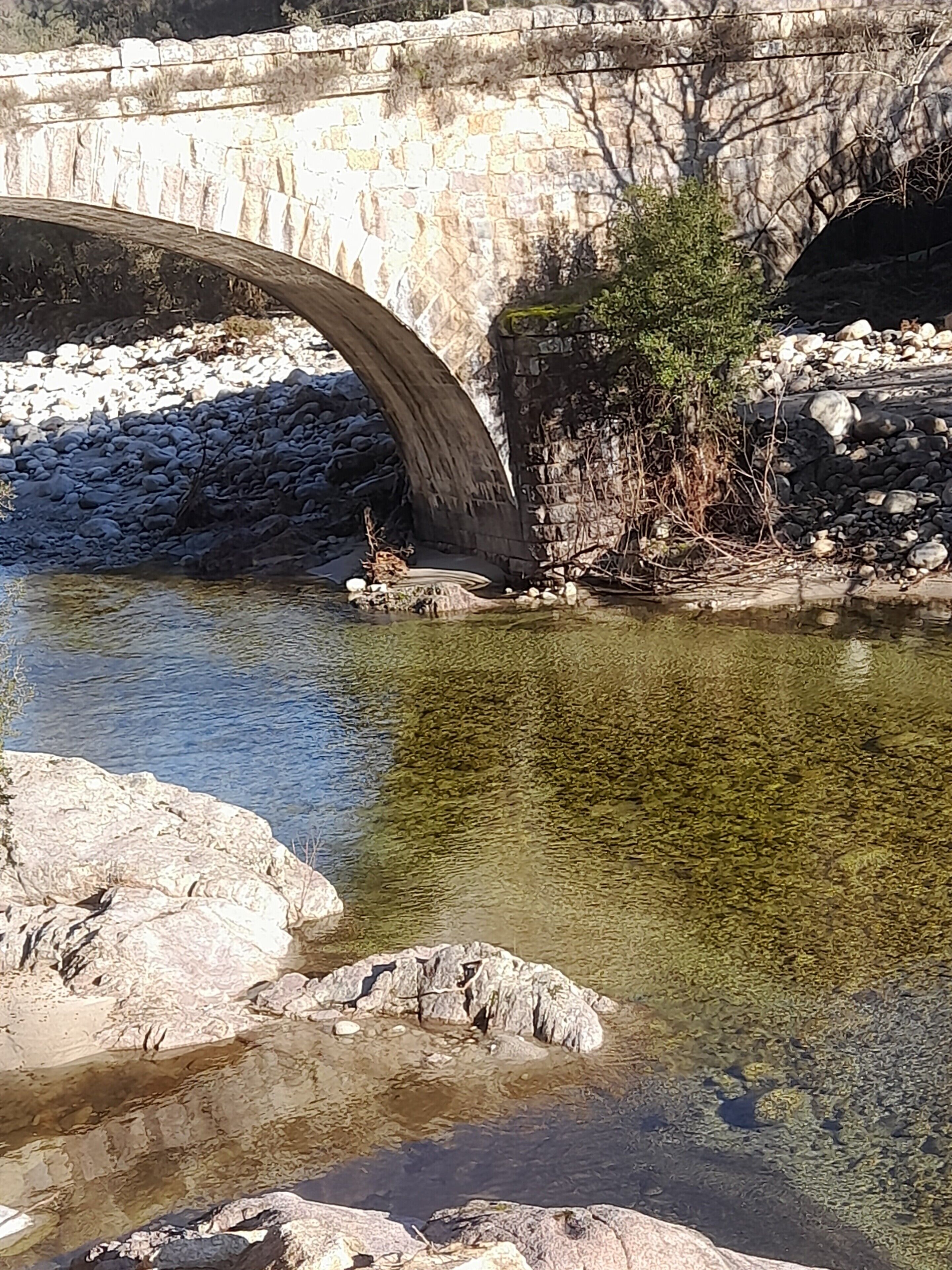 A stone bridge over a clear river with rocky banks and sparse vegetation.