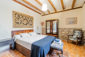 Un dormitorio con cama de madera, colcha azul, pared de piedra, armario de madera y una silla.
