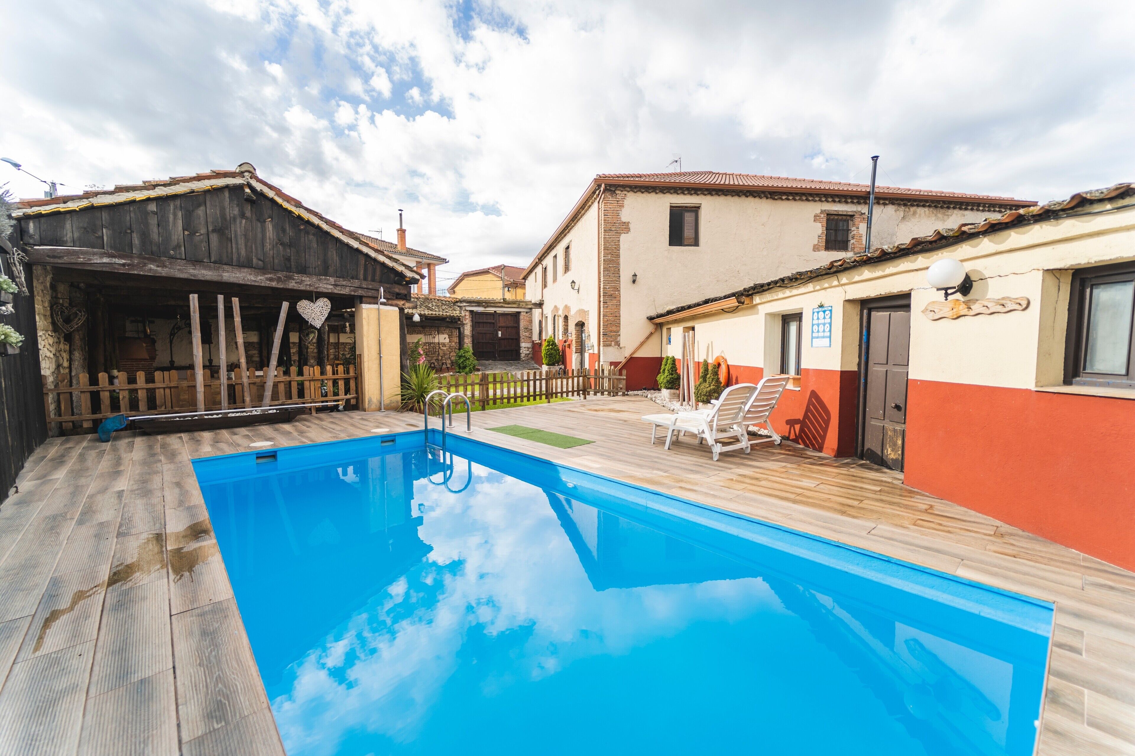 Une piscine avec une terrasse en bois, un espace délimité par une clôture et un bâtiment à la façade rouge et beige.