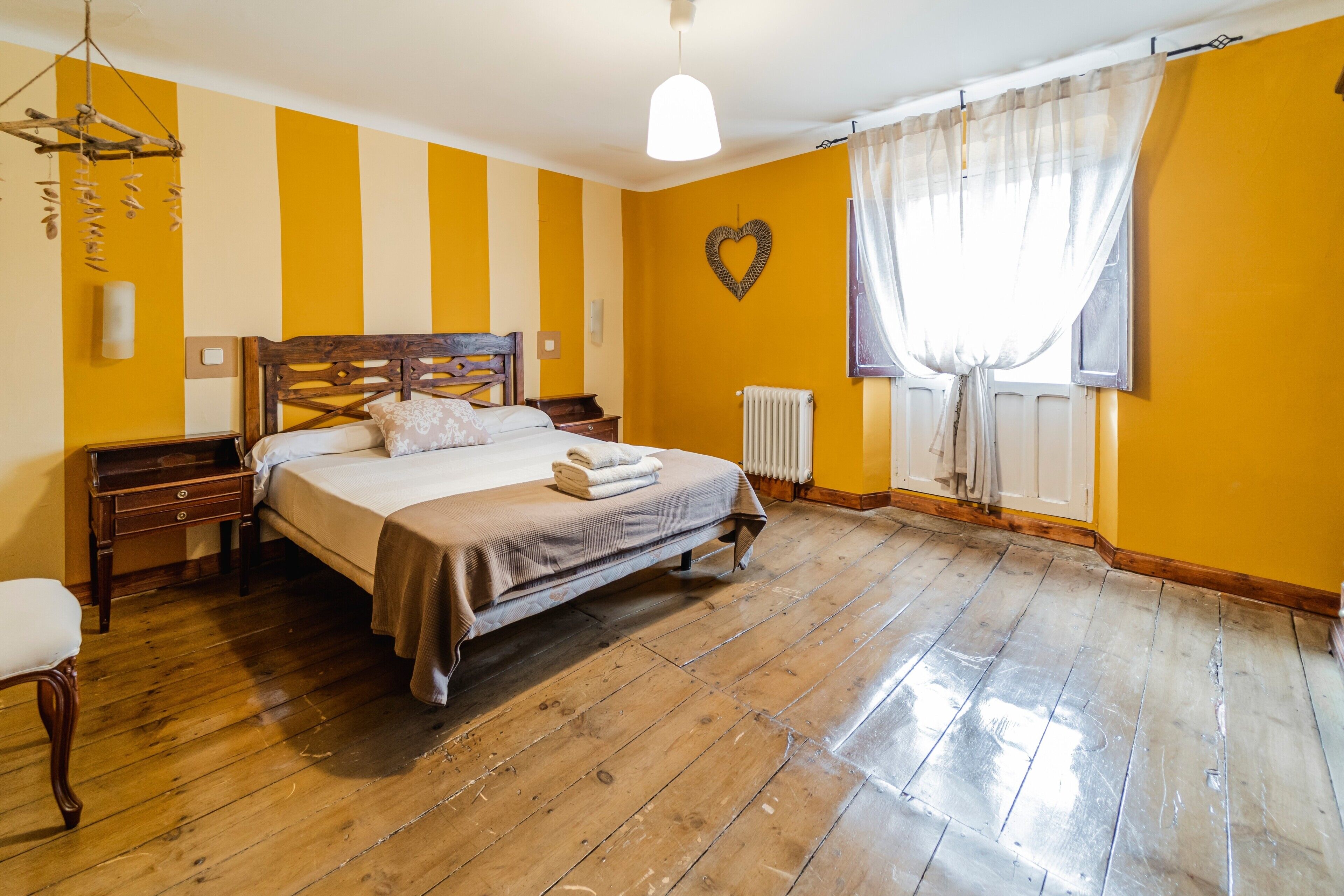 Un dormitorio con suelo de madera, una cama con cabecera, una mesita de noche, una silla, una lámpara colgante y una ventana con cortinas.