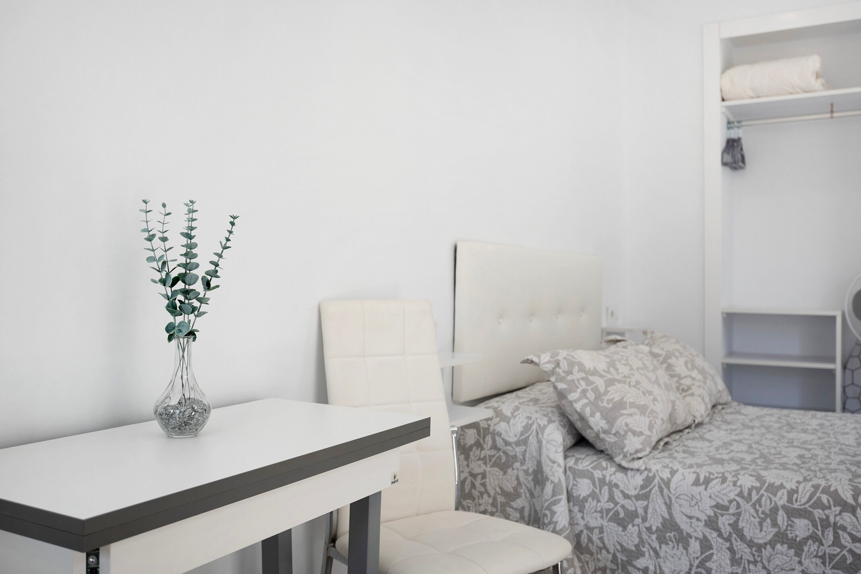Een slaapkamer met een bed, een nachtkastje, een stoel, een kledingkast en een vaas met een plant.