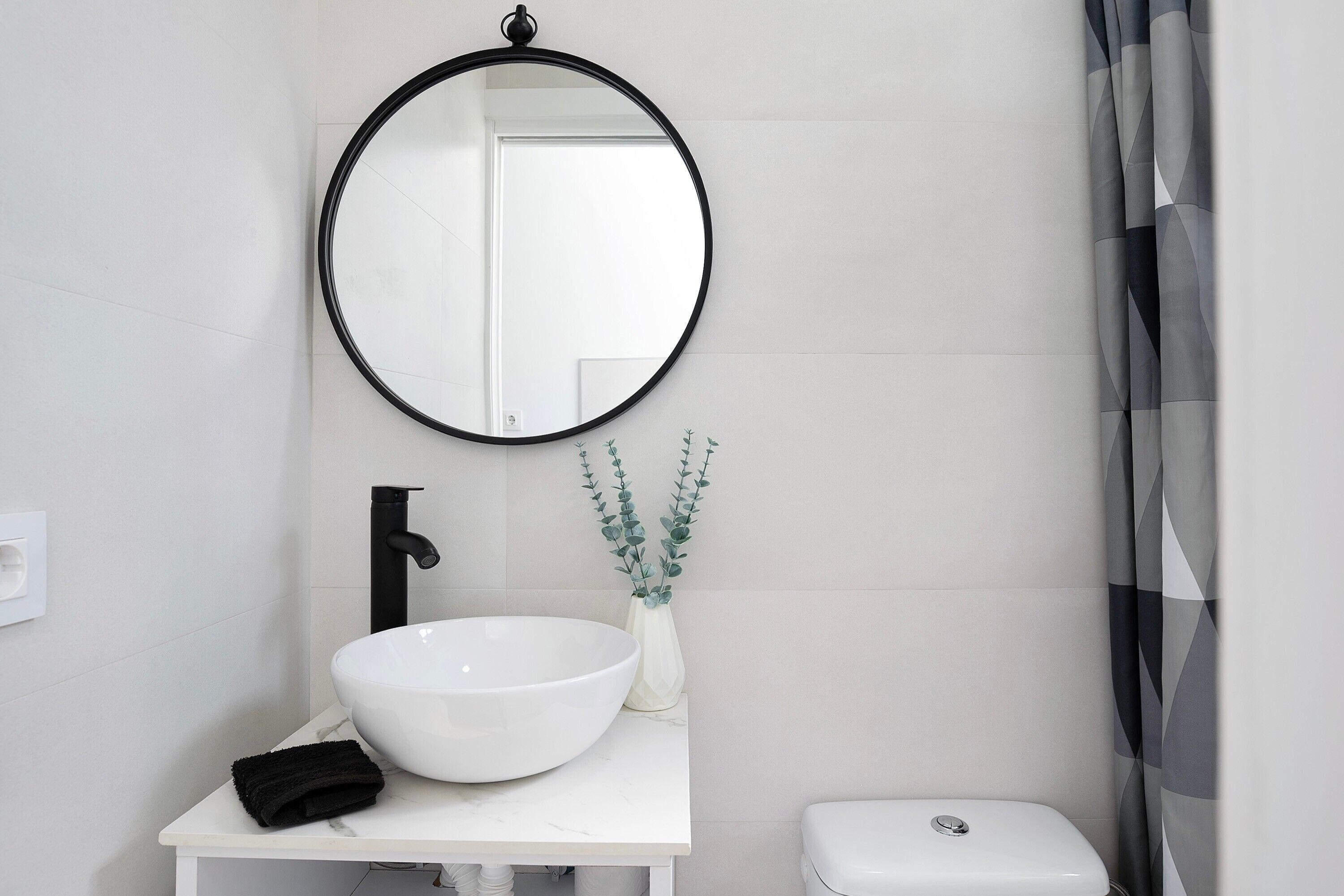 Un baño con lavamanos blanco, grifo negro, espejo redondo y retrete blanco.