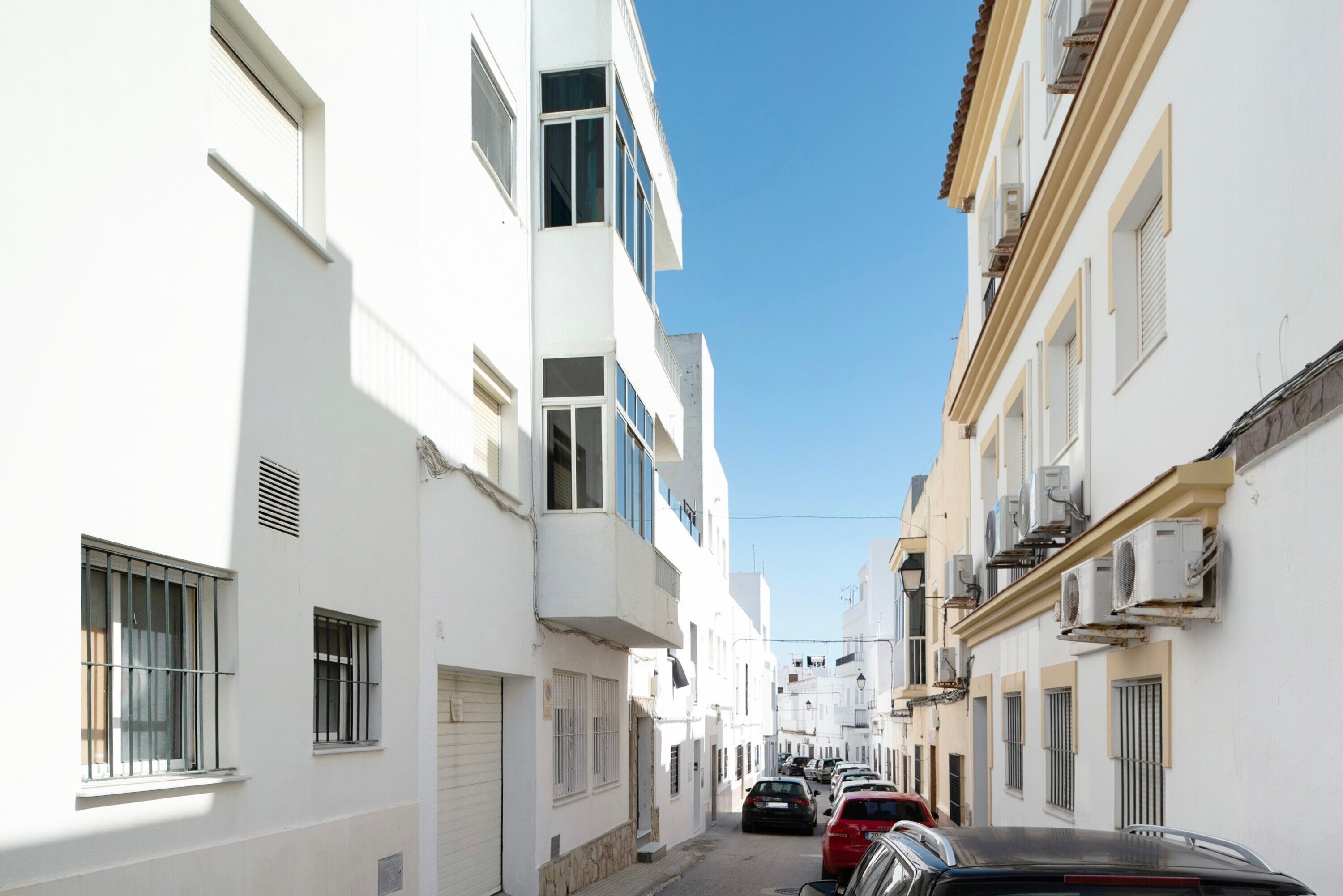 Un carrer estret amb edificis blancs, cotxes aparcats i un cel blau clar.