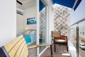 Un balcon avec une chaise en bois, un lit recouvert d’une couverture bleue et une plante fixée au mur.