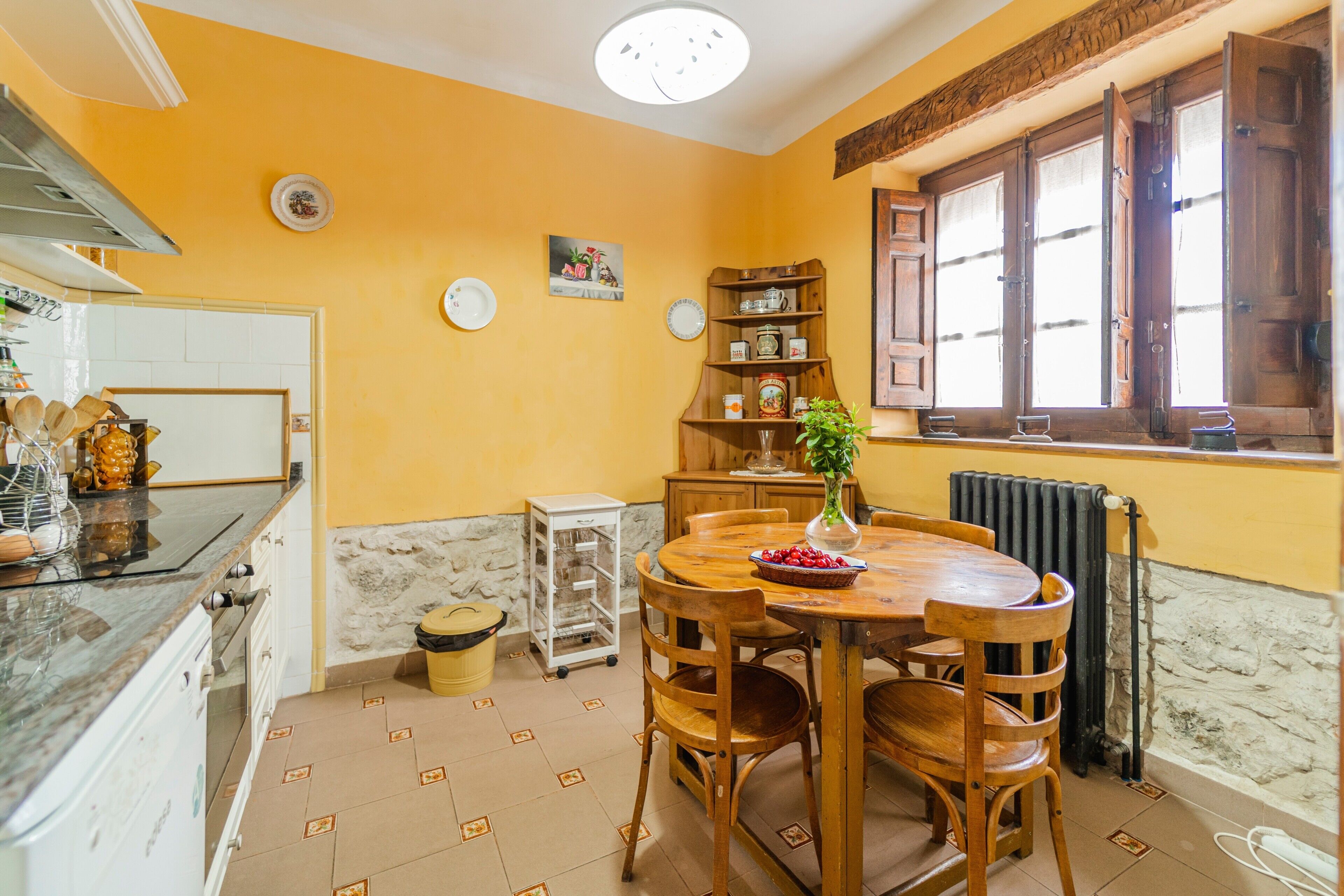 A kitchen with a dining table, chairs, a vase with flowers, a radiator, and a window with wooden shutters.