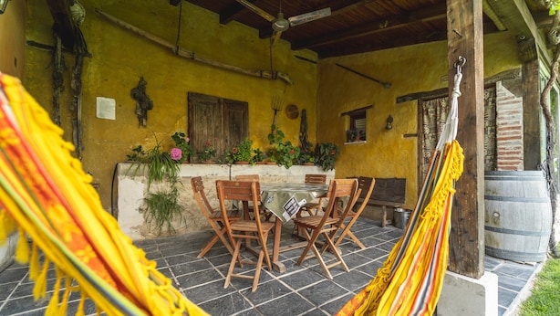 Un'amaca, sedie di legno, un tavolo e un muro rustico giallo con piante.