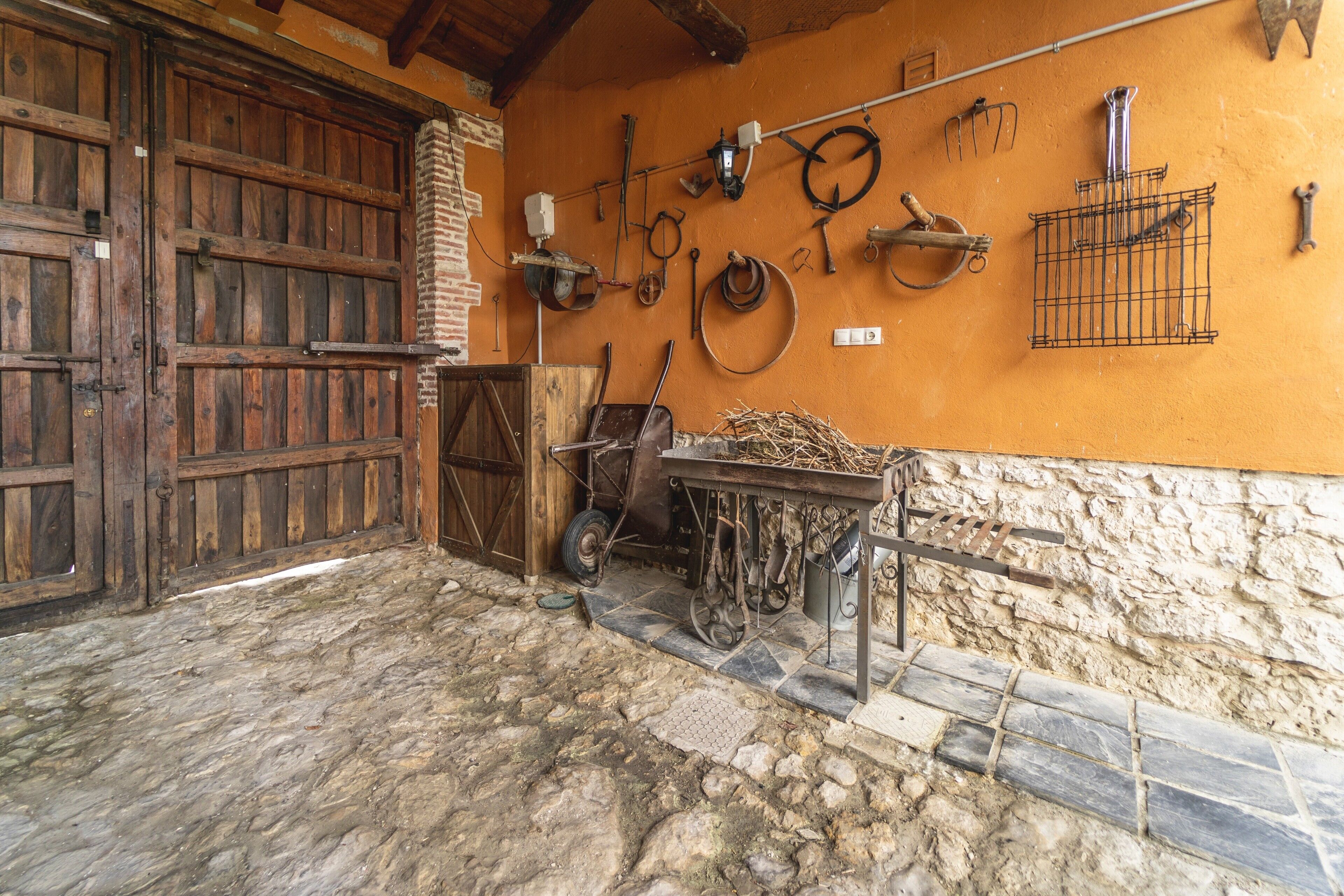 Een rustiek ogende schuur met houten deur, stenen vloer en diverse landbouwgereedschappen en -materieel aan de wand bevestigd.