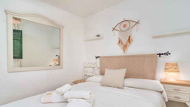 Ein ordentlich bezogenes Bett mit weißen Leinen, einer Nachttischlampe und einem an der Wand befestigten Traumfänger.