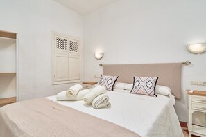A neatly made bed with rolled towels, a headboard, two bedside tables, a wardrobe, and a wall-mounted light fixture.